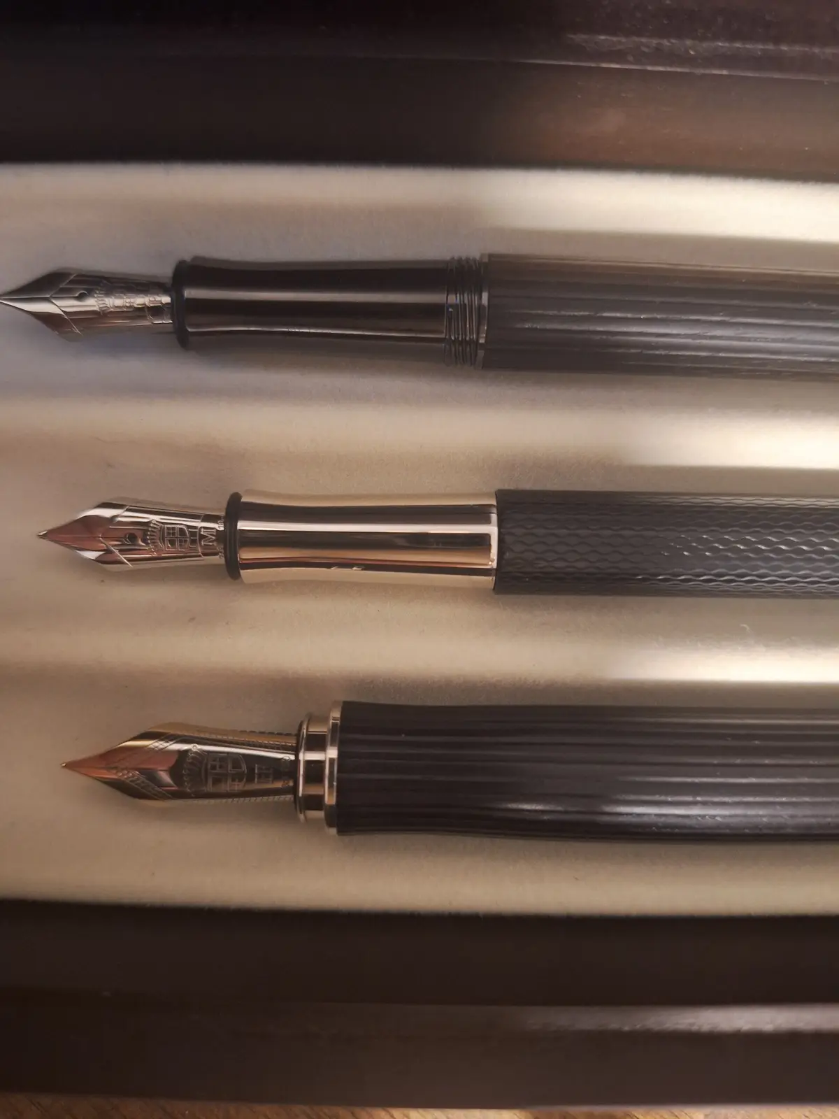 Three identical luxury fountain pens are laid horizontally in a row on a light-colored, textured surface, each with a…