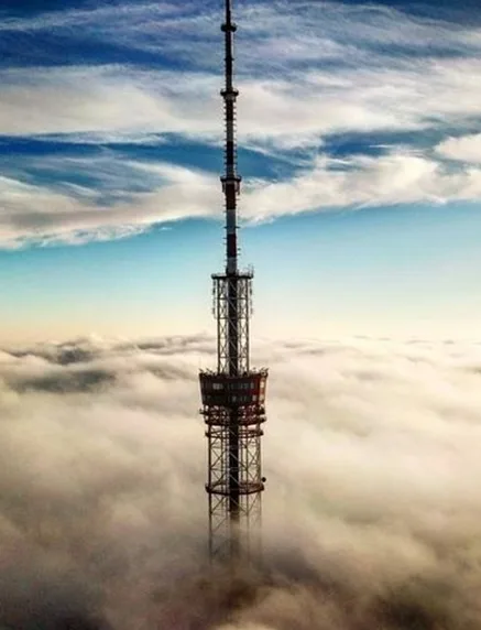 A tall metal broadcast tower stands piercing through a thick layer of clouds, with a blue sky and wispy clouds above. The…