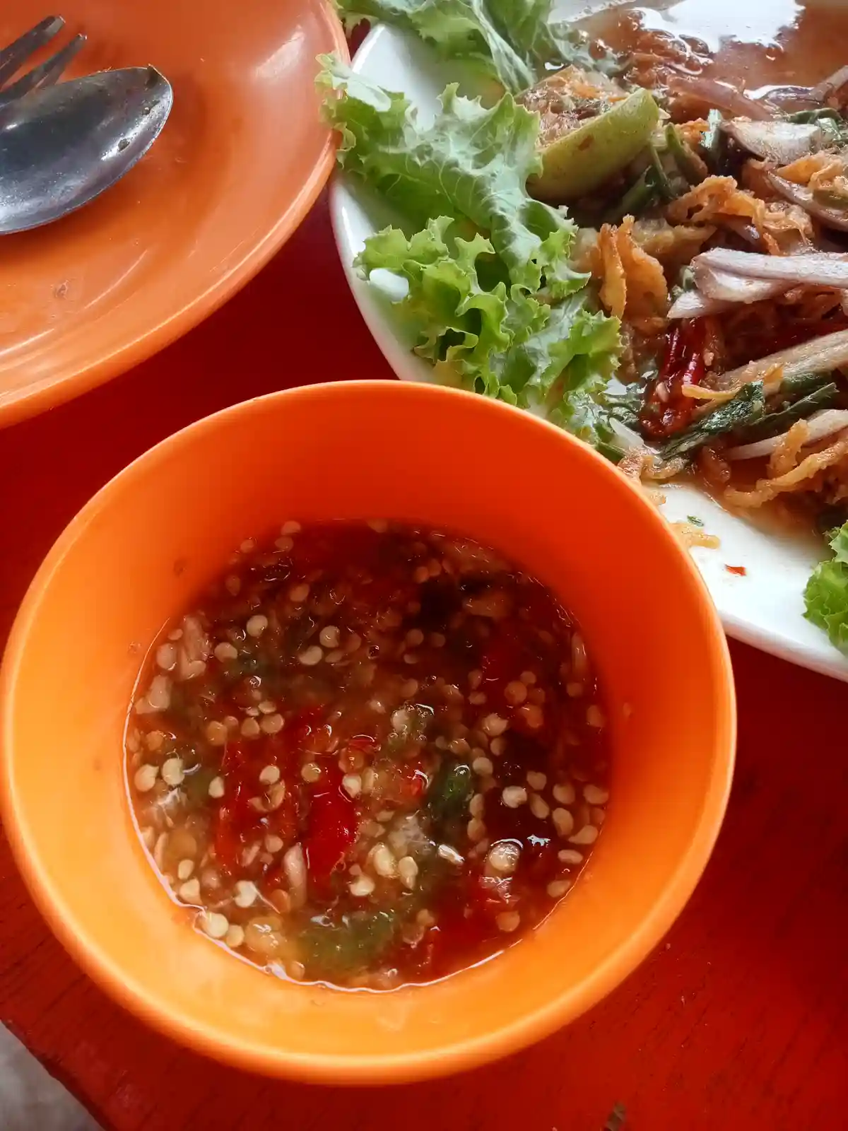 A close-up photo shows a bright orange plastic bowl filled with a reddish, chunky sauce containing visible red chili pieces…