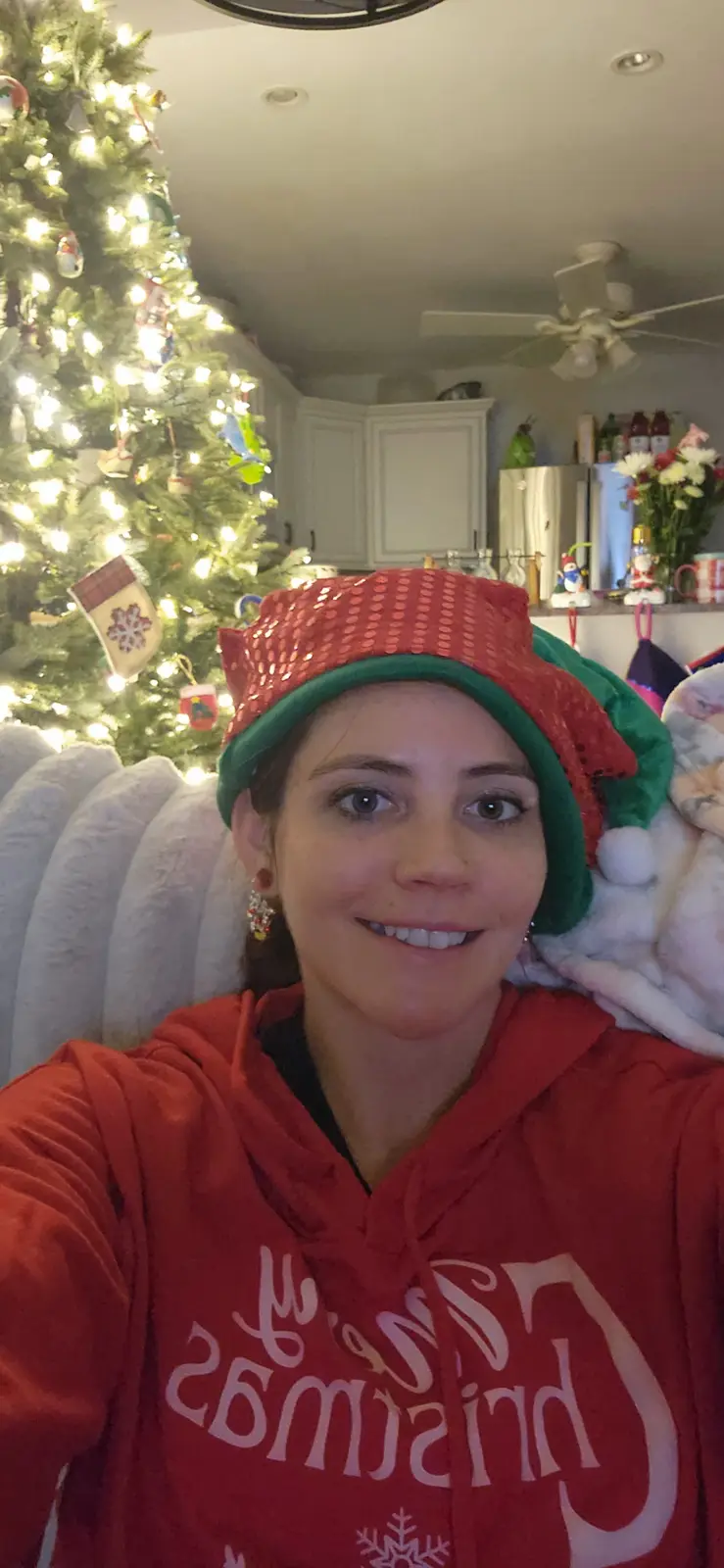 A woman in a red and green elf hat and a red 'Merry Christmas' hoodie smiles at the camera while sitting in front of a lit…