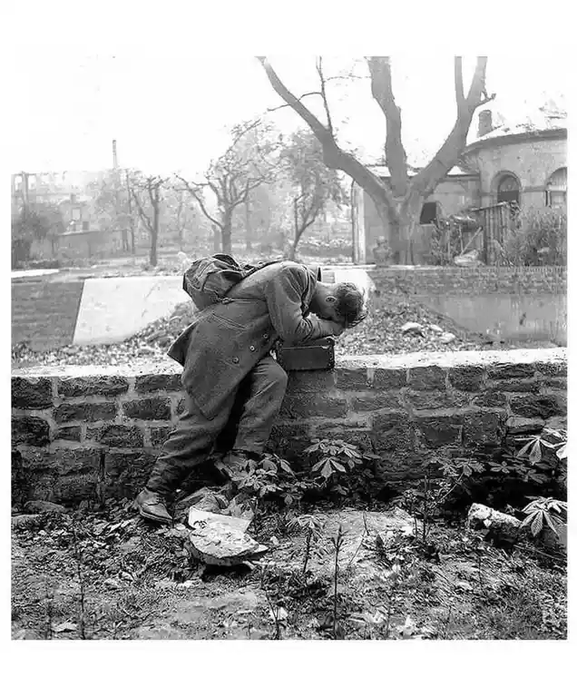 A black-and-white historical photo shows a man in a trench coat and backpack leaning heavily against a stone wall, his head…