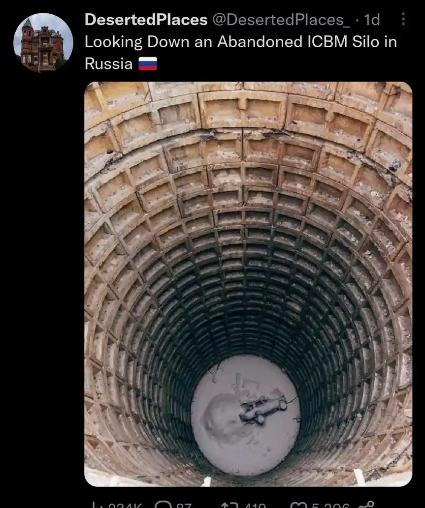 A top-down view into a massive, circular, abandoned concrete silo with deteriorating walls and a small, overturned car at…