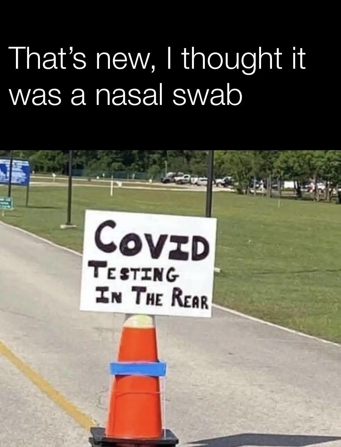 An orange traffic cone with a handwritten sign that reads 'COVID TESTING IN THE REAR' is placed on a paved road next to a…