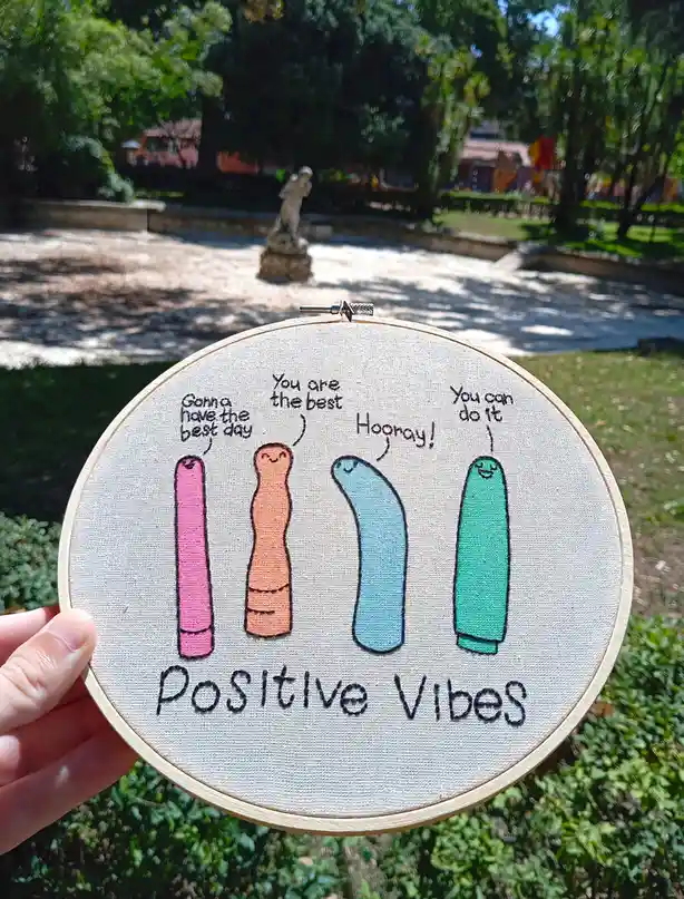 A hand holds a circular embroidery hoop displaying four colorful, smiling, finger-like characters. Each character has a…