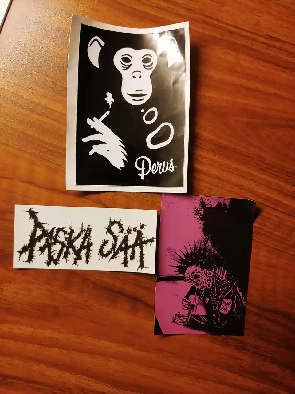 Three stickers lie on a wooden surface: a black-and-white one of a monkey smoking with 'Perus' written below, a white one…