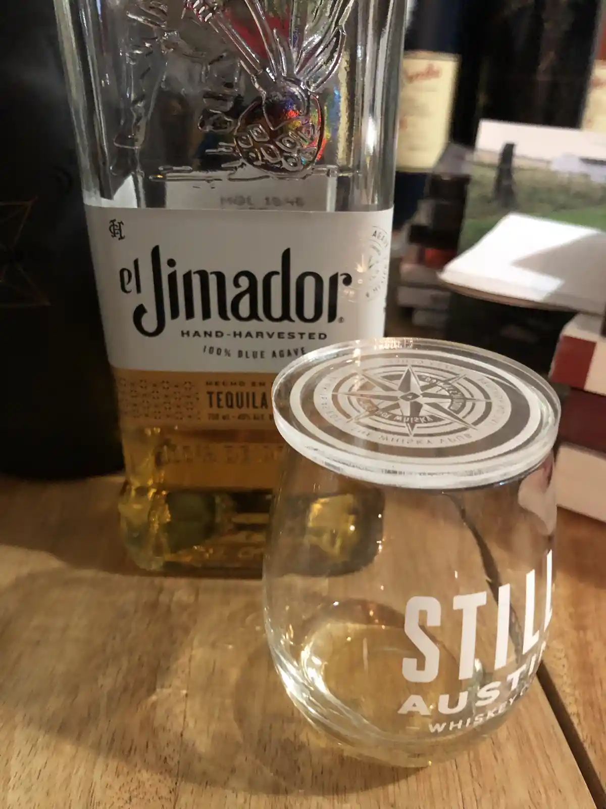 A clear glass tumbler with a compass rose lid and 'STILL AUSTIN WHISKEY' printed on it sits on a wooden table next to a…