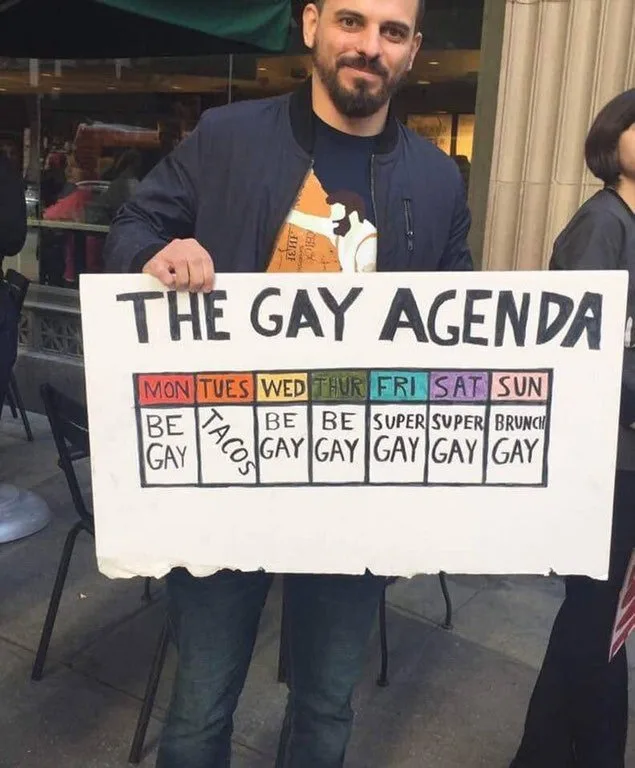 A man holds a handmade white sign titled 'THE GAY AGENDA' featuring a weekly schedule with days of the week and activities…