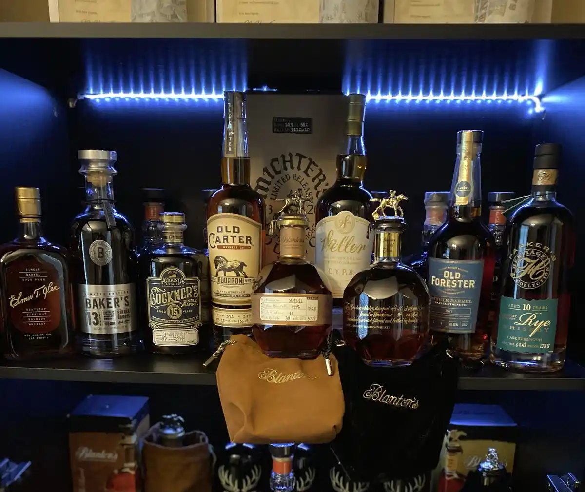 A well-lit shelf displays numerous expensive bourbon and rye whiskey bottles, including Blanton's, Baker's, and Old…