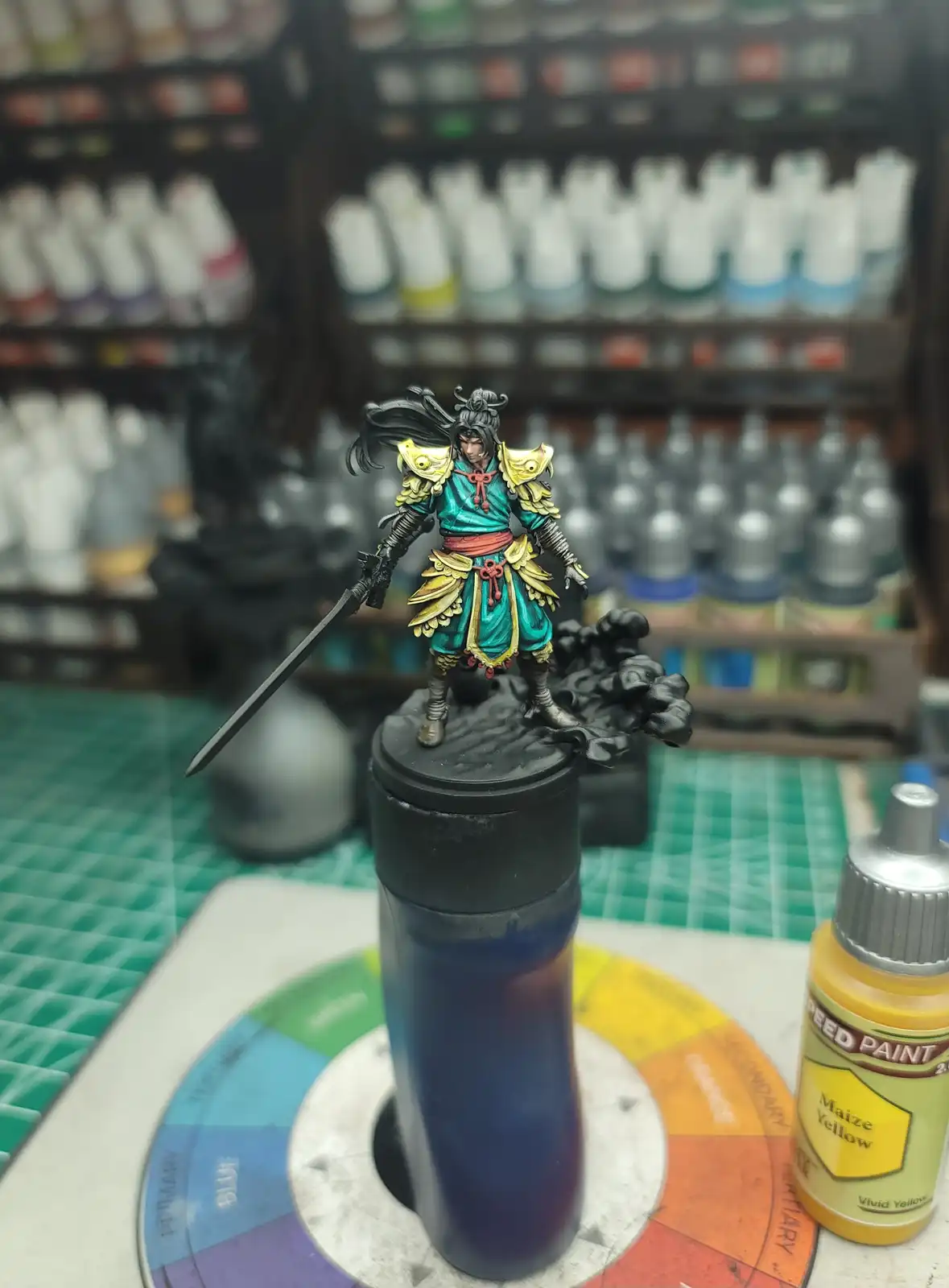 A highly detailed, painted miniature warrior figure with teal and gold armor stands atop a black paint bottle, holding a…