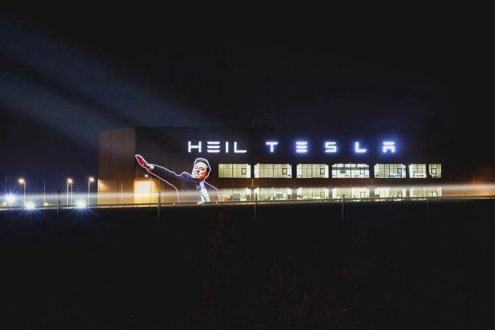 A nighttime photo of a large building with a projection of a man in a suit raising his right hand, above which the words…