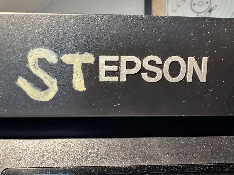 A close-up of a black Epson device shows the brand name 'EPSON' in silver letters, but the 'E' is replaced with a crudely…