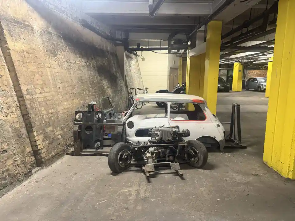 A partially disassembled white Mini car sits in an underground parking garage, its body shell separated from the chassis and…