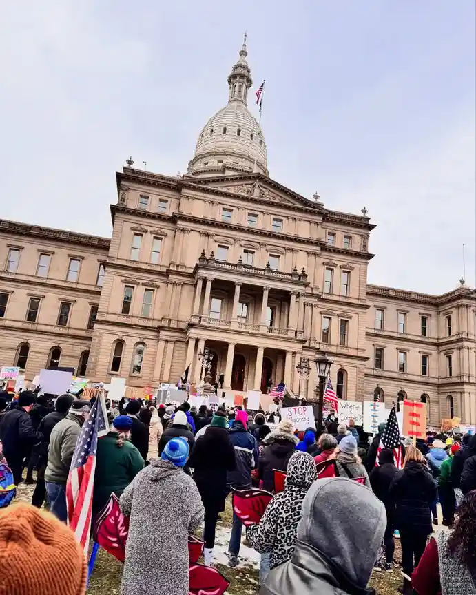 A large crowd of protesters stands in front of a grand, domed state capitol building, holding signs and American flags…