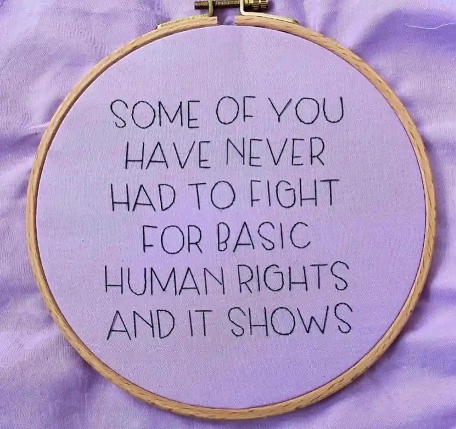 A close-up photo shows a light purple fabric stretched taut in a wooden embroidery hoop. Black thread is stitched in a…