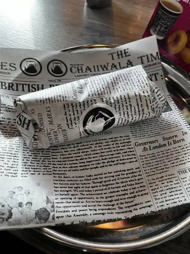 A rolled-up food item, likely a kebab, is wrapped in newspaper printed with 'THE CHAIWALA TIMES' and headlines like…