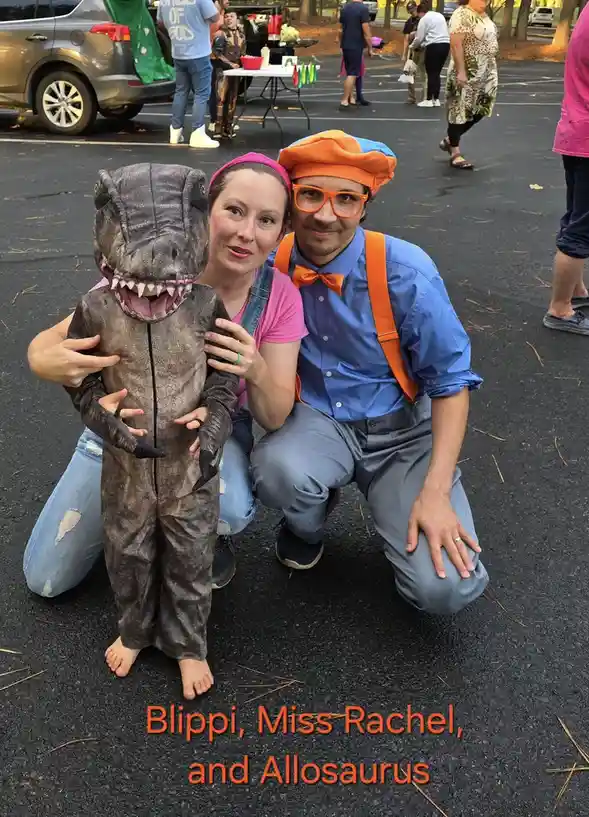 A photo of a man dressed as Blippi (blue shirt, orange bowtie, suspenders, and large orange glasses) kneeling next to a…