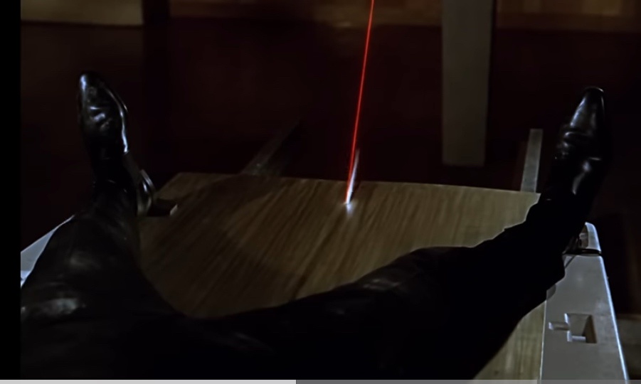 A person in black leather pants and shoes has their legs propped up on a wooden table, with a red laser beam shining…