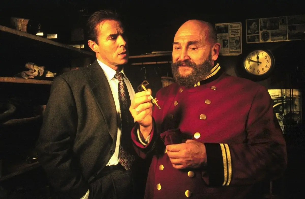 Two men stand in a dimly lit room; one in a suit holds a key and looks concerned, while the other in a red uniform with gold…