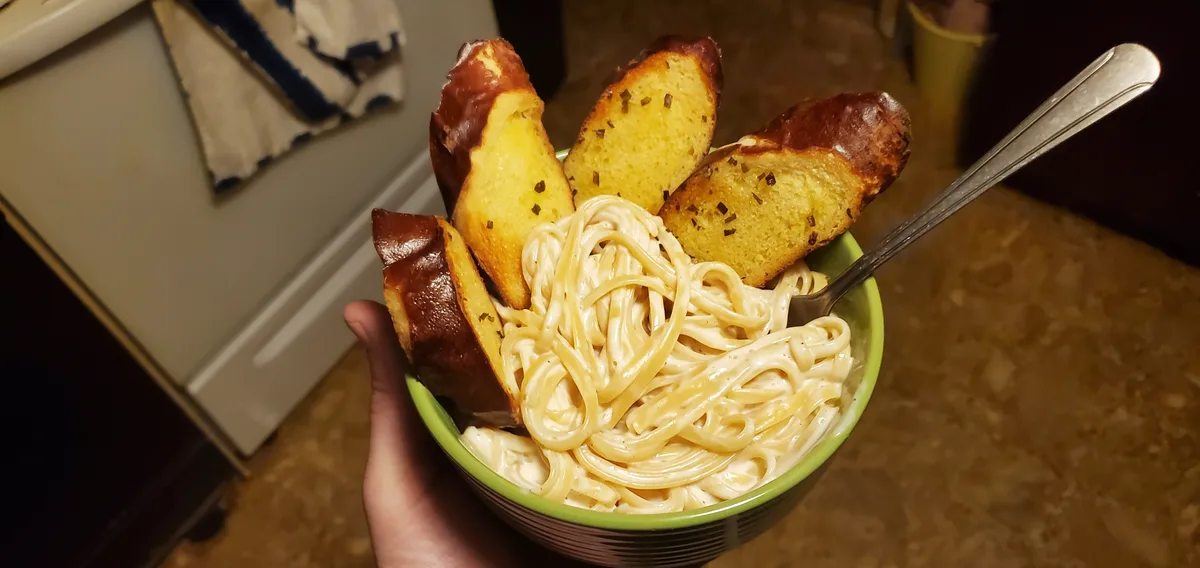 A hand holds a green bowl filled with creamy pasta and four slices of toasted garlic bread arranged around the edges. A…