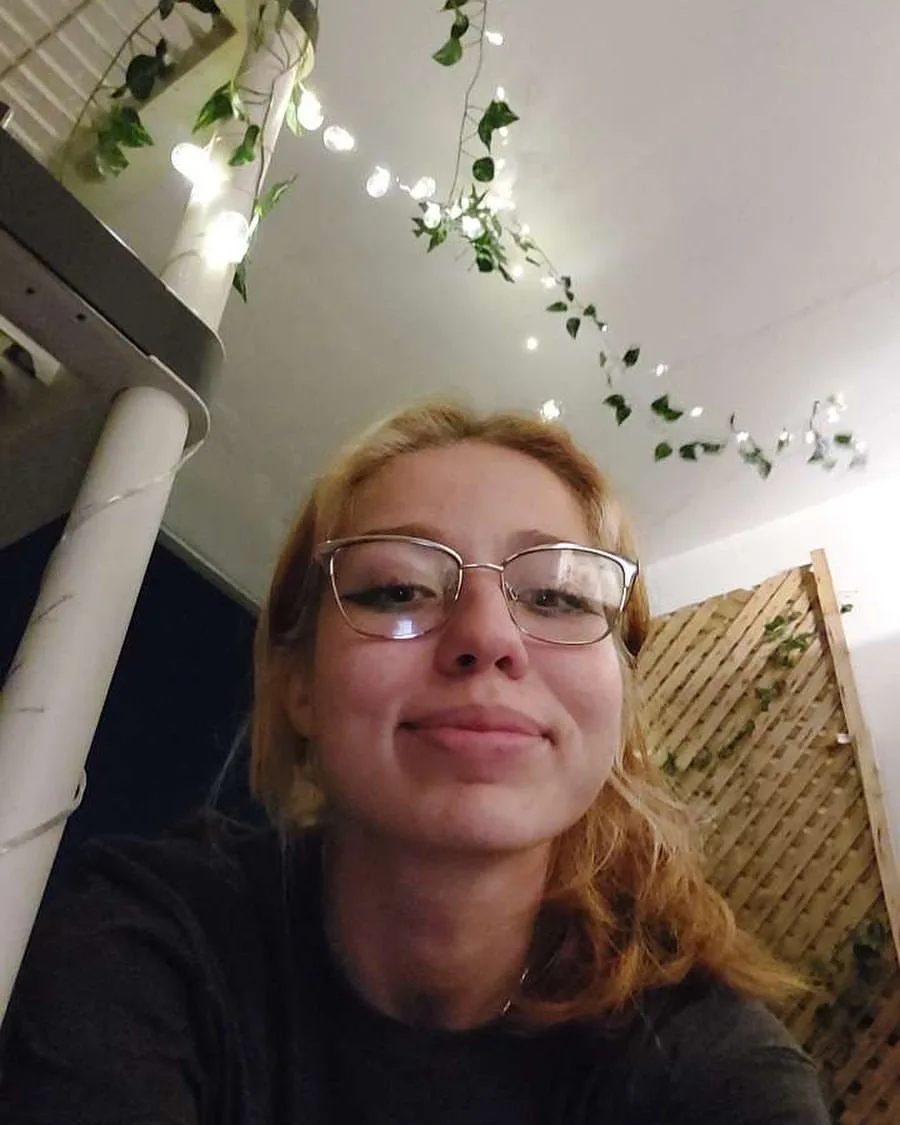 A selfie of a young woman with glasses and reddish-blonde hair, smiling slightly while looking at the camera. The background…