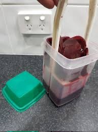 A hand uses wooden chopsticks to lift a dark red, gelatinous-looking food item from a clear plastic container, with the…