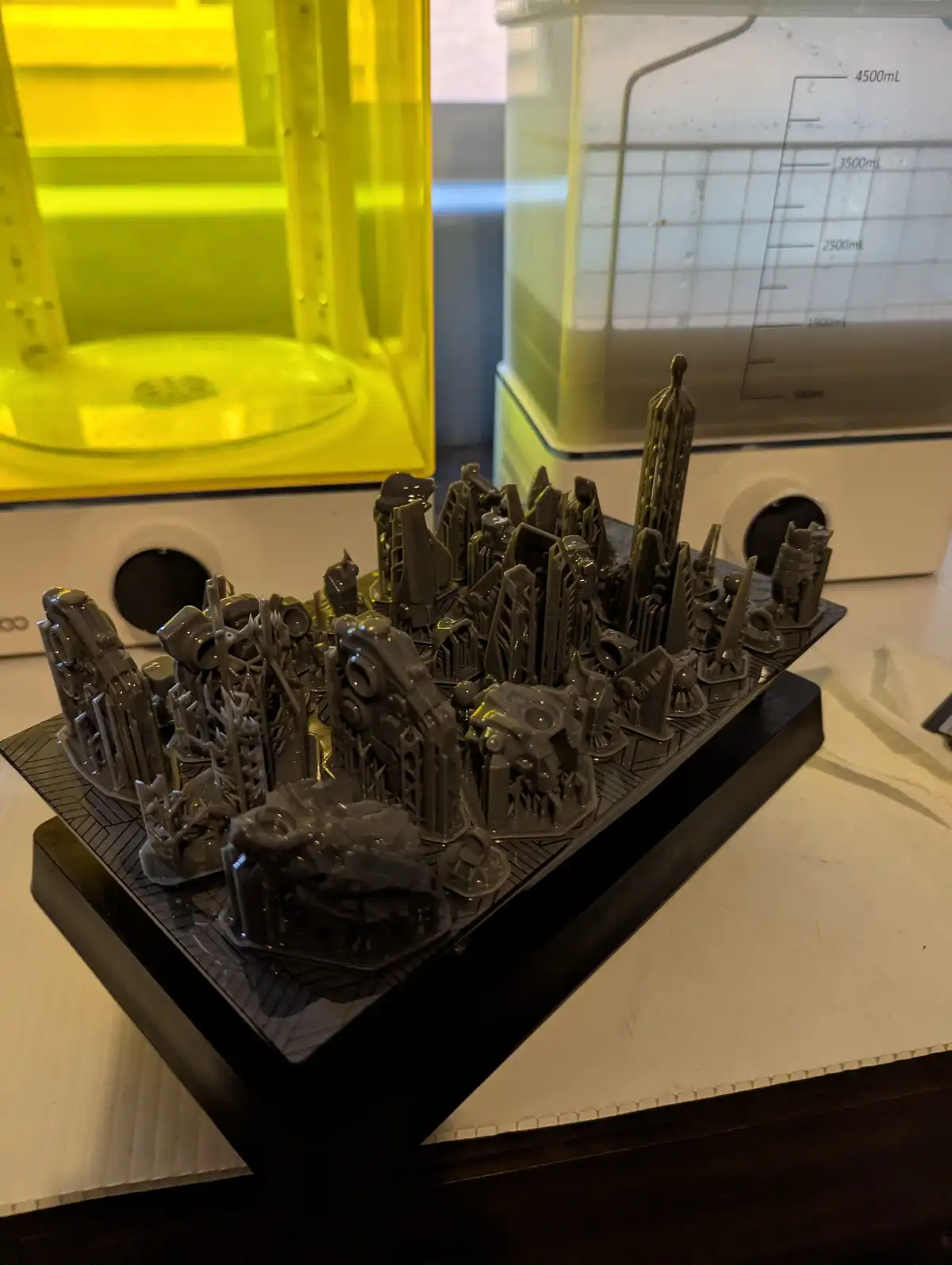 A 3D-printed miniature cityscape with intricate, futuristic buildings sits on a black base, placed on a white countertop…