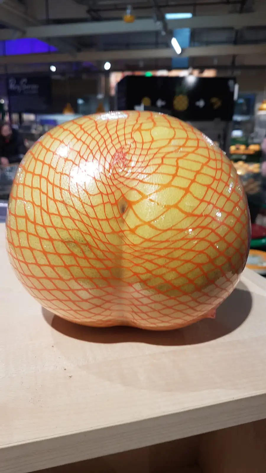A close-up photo shows a large, round citrus fruit, likely a grapefruit, resting on a light-colored wooden surface in what…