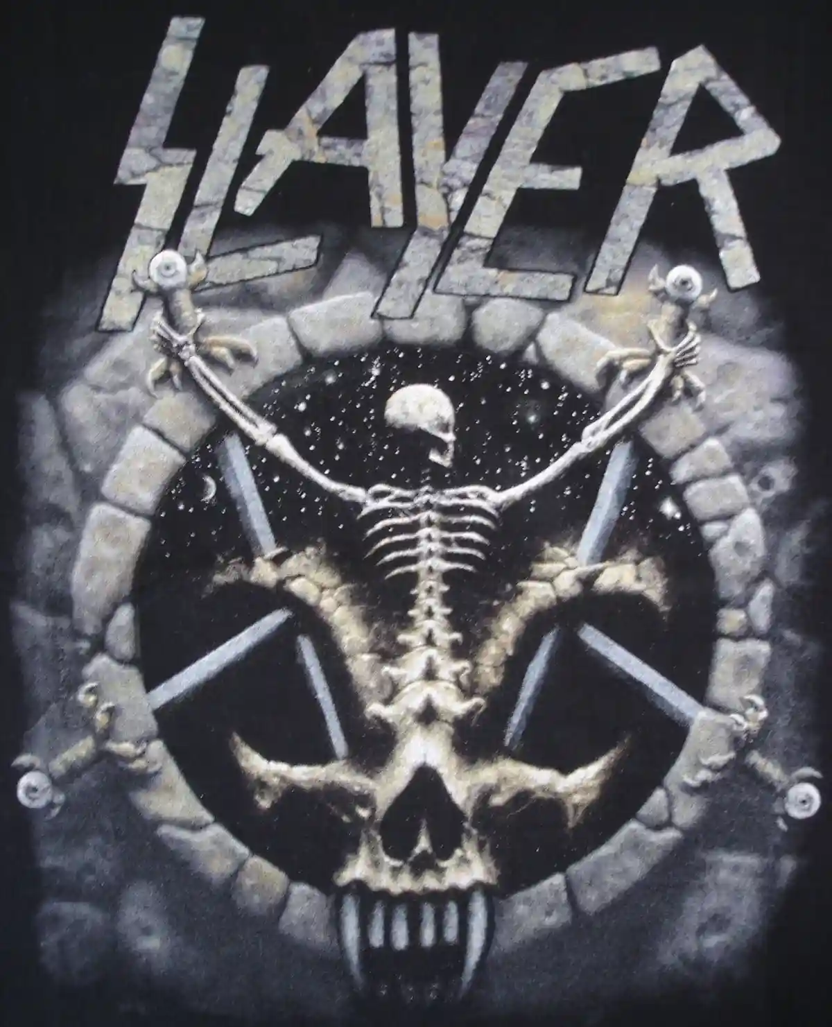 A black t-shirt features the iconic Slayer band logo in a stone-like font above a circular stone archway. Inside the arch, a…