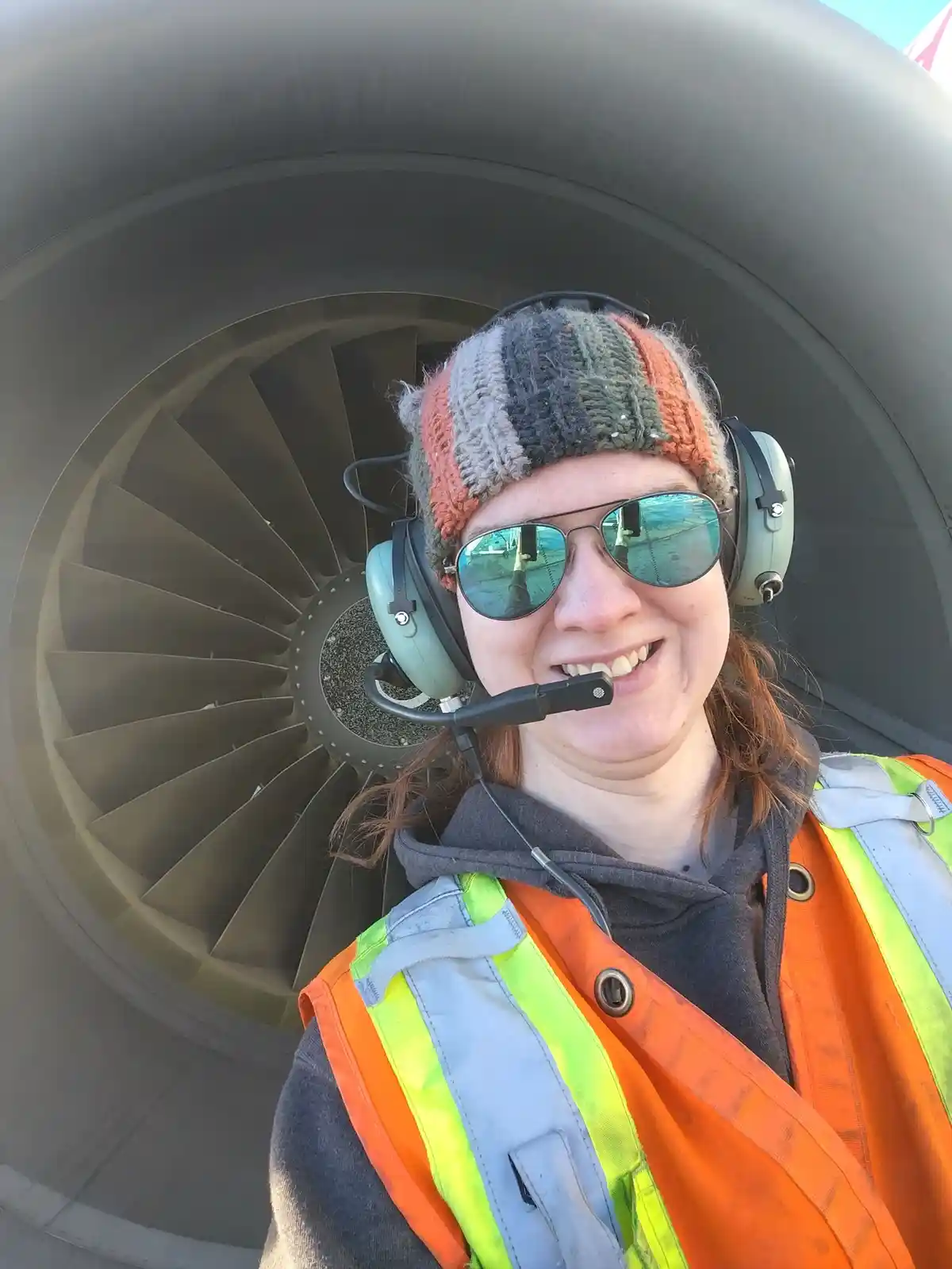 A smiling person wearing a striped knit hat, aviator sunglasses, and a headset with a microphone, stands in front of a large…