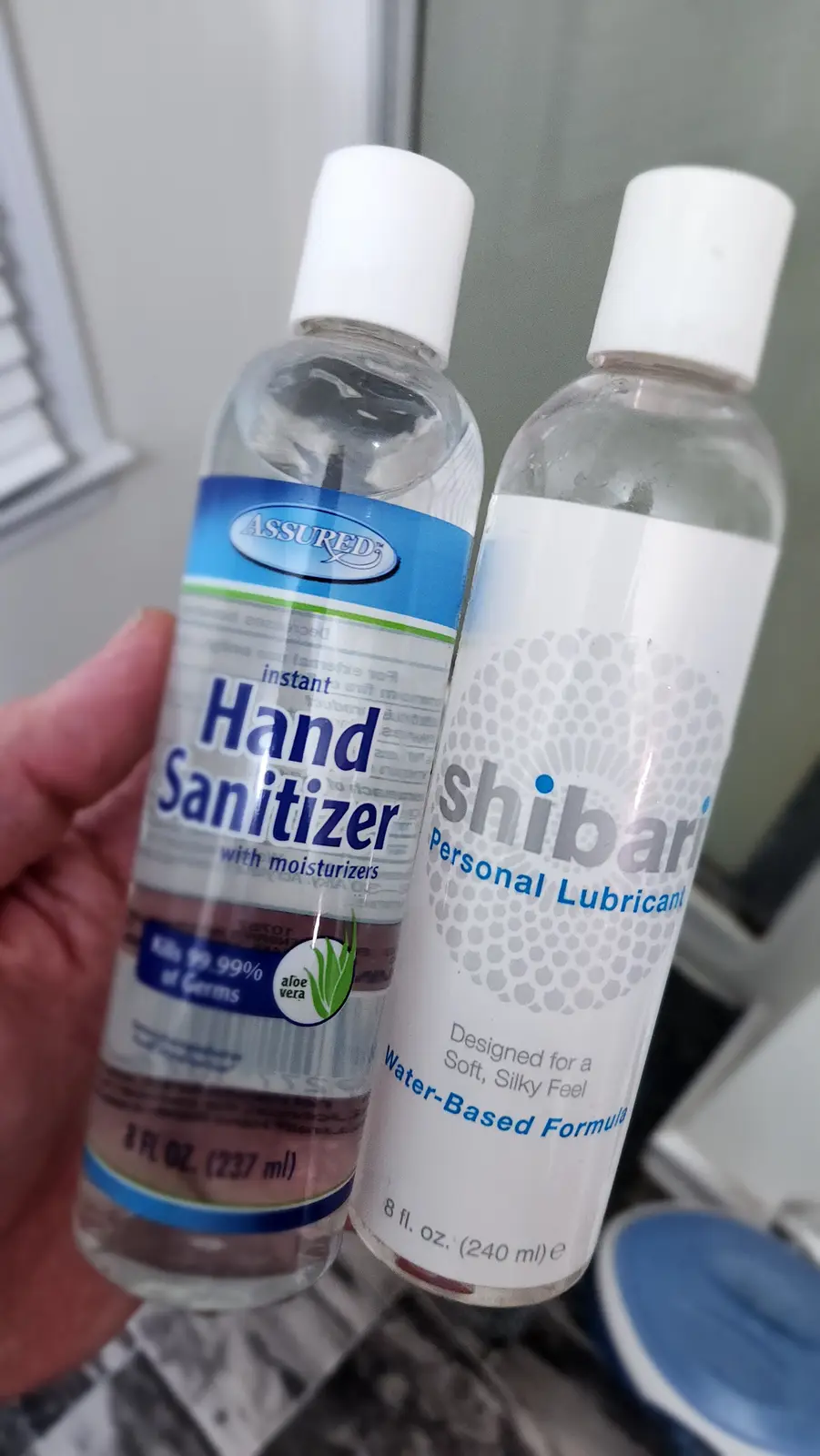 A hand holding two clear plastic bottles side by side: one labeled 'Assured instant Hand Sanitizer with moisturizers' and…