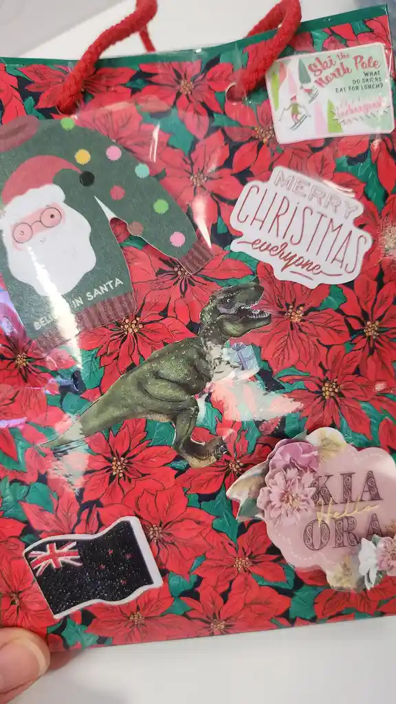 A Christmas gift bag covered in red poinsettia flowers has multiple stickers applied to it, including a cartoon Santa in a…
