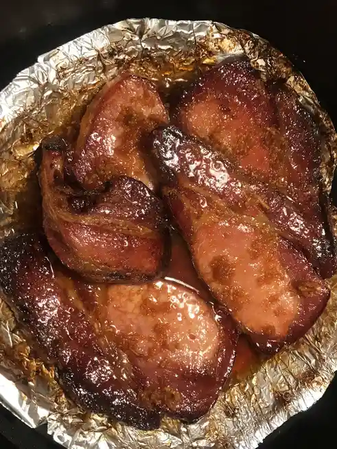 A close-up photo shows several slices of cooked bacon resting on crumpled aluminum foil inside a dark pan. The bacon is…