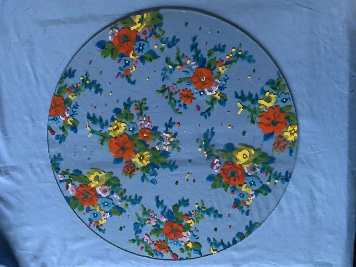 A circular glass plate with thick, colorful, textured paint depicting flowers in red, orange, yellow, blue, and green, set…