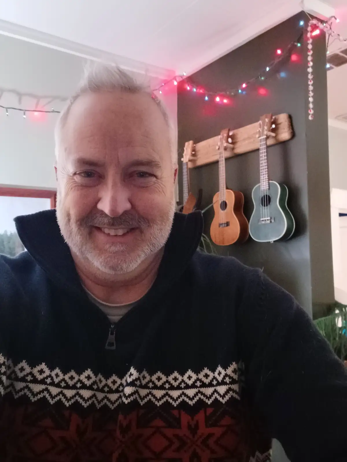 A smiling middle-aged man with a gray beard and hair wears a dark zip-up sweater with a red and white patterned yoke. Behind…
