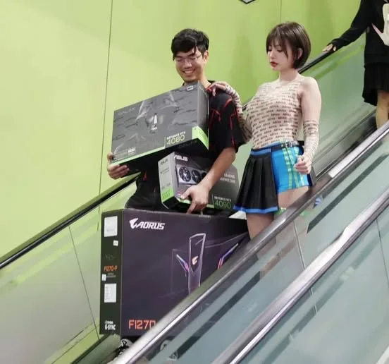 A young man and woman are riding an escalator, the man is carrying several large computer hardware boxes, including AORUS…