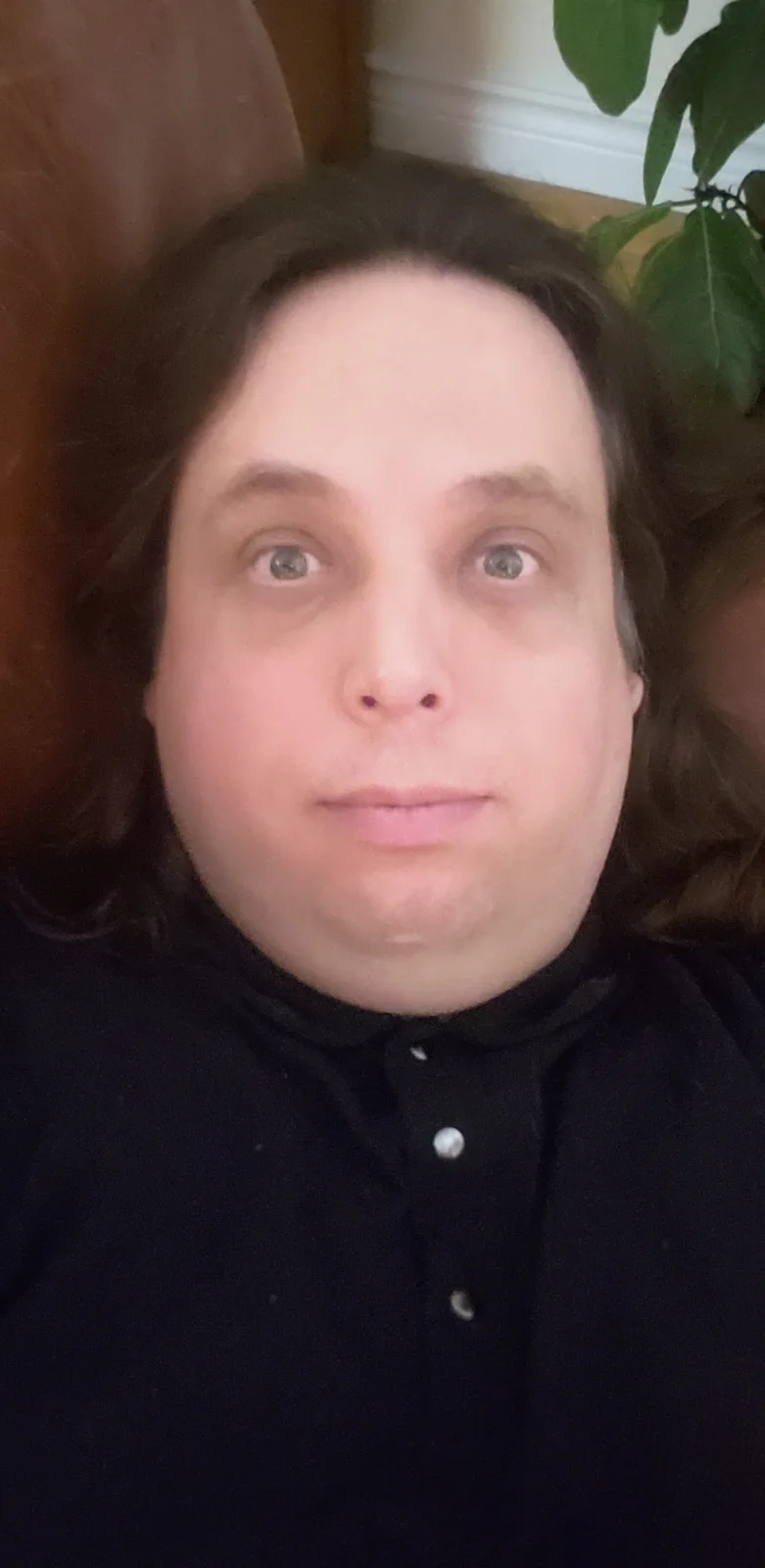 A close-up selfie of a person with long brown hair and a black button-up shirt, looking directly at the camera with a blank…