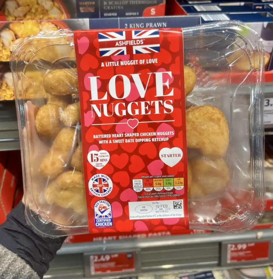 A hand holds a clear plastic package of heart-shaped chicken nuggets in a supermarket aisle. The red label reads 'ASHFIELDS'…