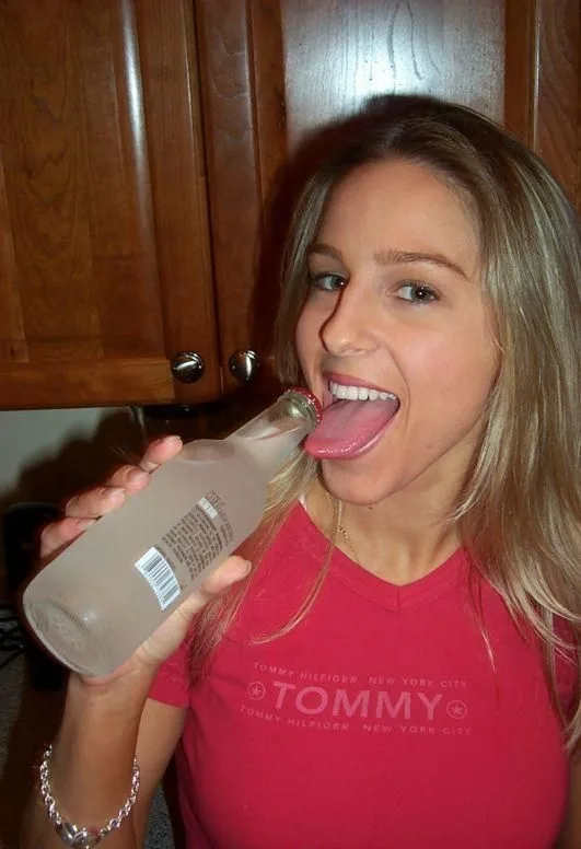 a young woman with blonde hair wearing a red Tommy Hilfiger t-shirt is holding a clear glass bottle with a red cap, sticking…