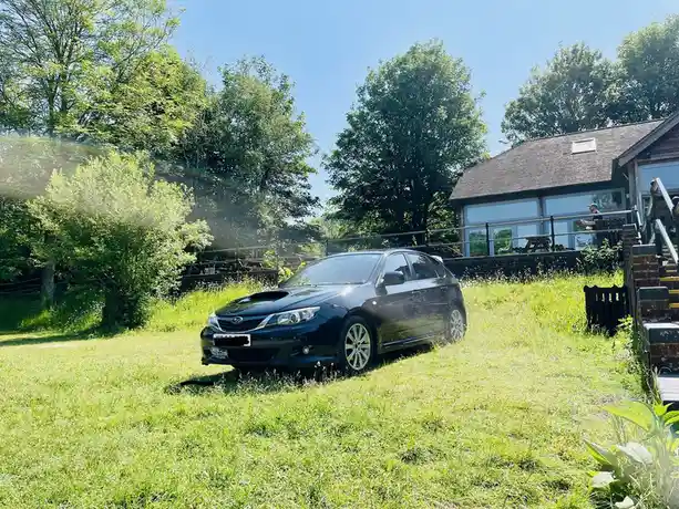 A black Subaru Impreza WRX STI is parked on a grassy lawn in front of a house with large windows and a wooden deck. The car…