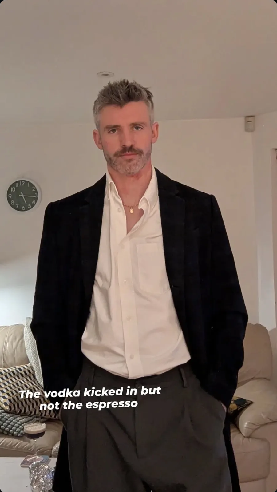 A man with gray hair and a beard, wearing a black blazer over a white shirt and dark pants, stands in a living room with his…