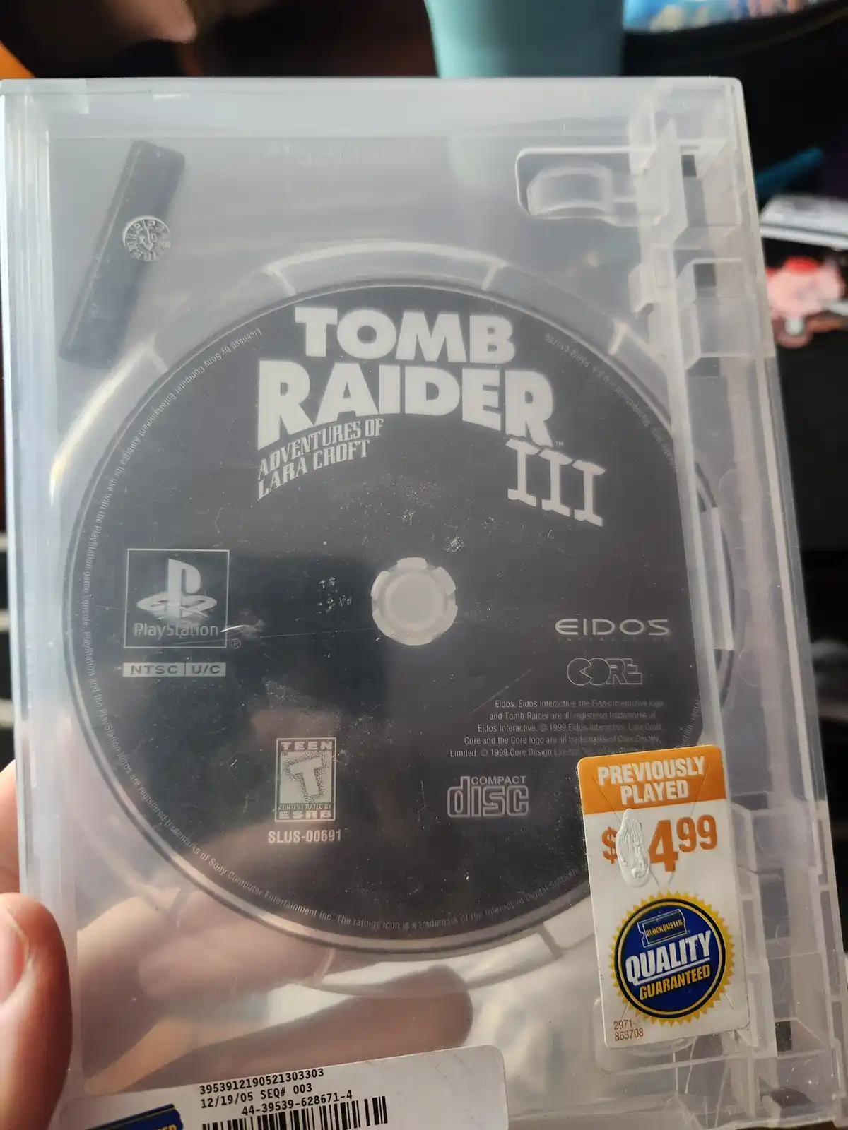A close-up photo of a PlayStation game disc for 'Tomb Raider III: Adventures of Lara Croft' inside its clear plastic case. A…