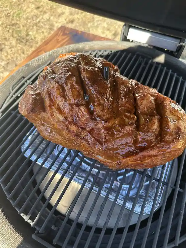 A large, heavily seasoned, and glistening steak sits on the grates of a grill, with a small black thermometer probe inserted…