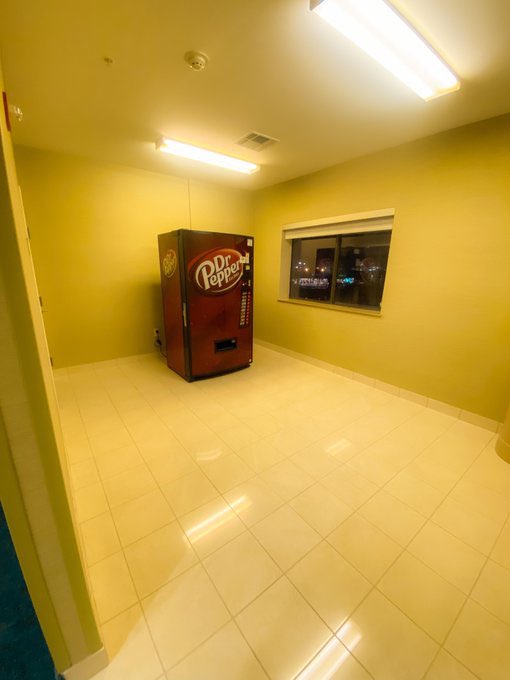 A red Dr Pepper vending machine stands alone in a brightly lit, empty room with yellow walls and white tiled floors, viewed…