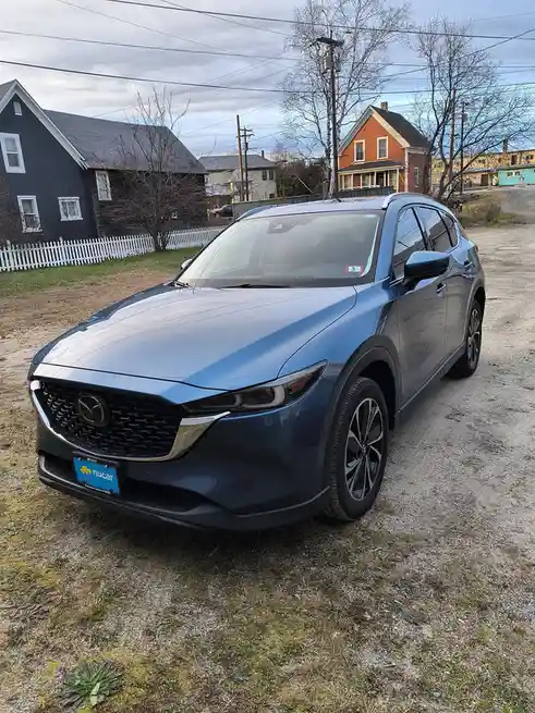 A blue Mazda CX-30 SUV is parked on a gravel driveway in front of residential houses with bare trees and power lines under…