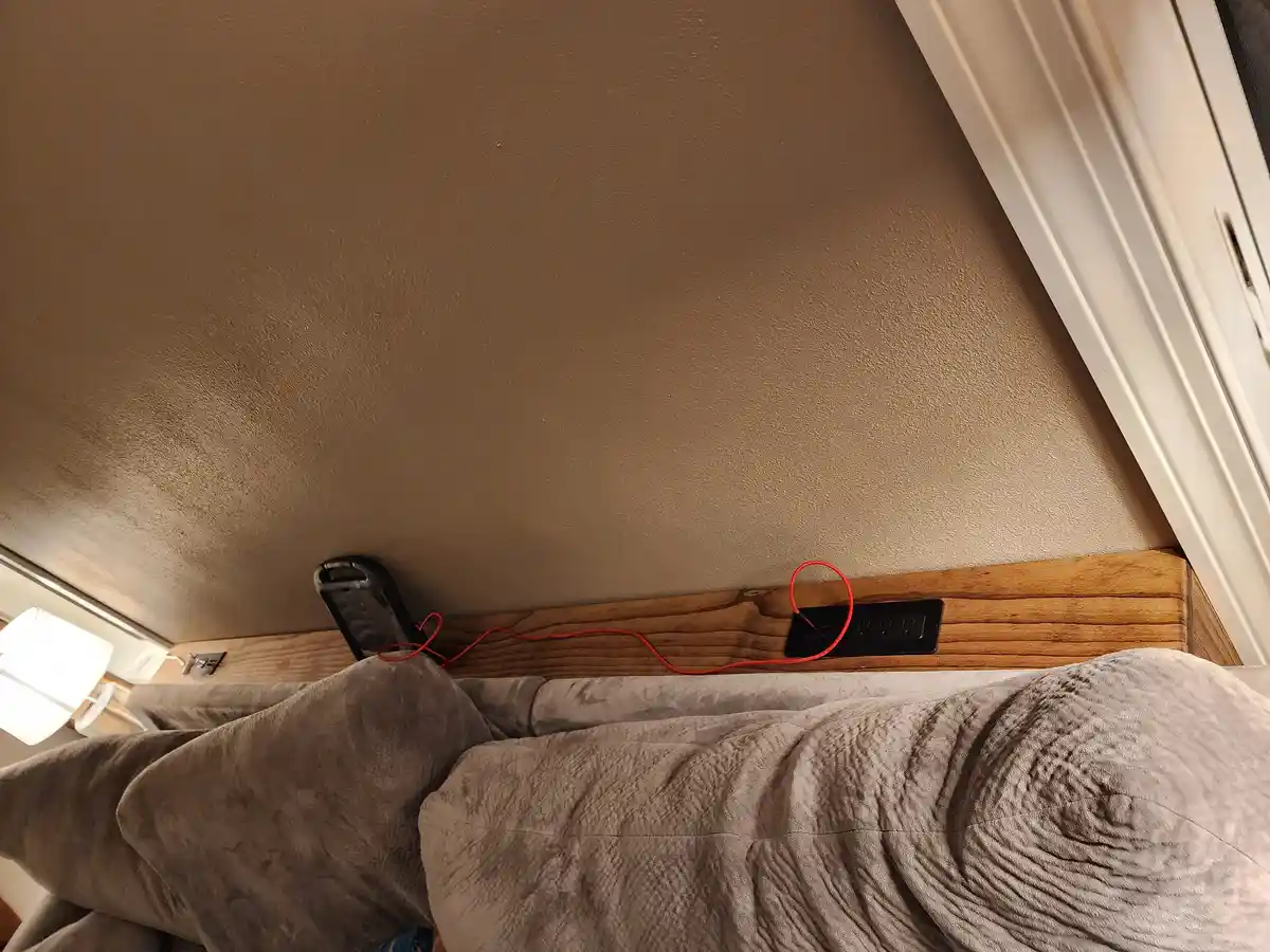 A tilted photo shows a red phone charging cable plugged into a black wall outlet mounted on a wooden headboard above a gray…