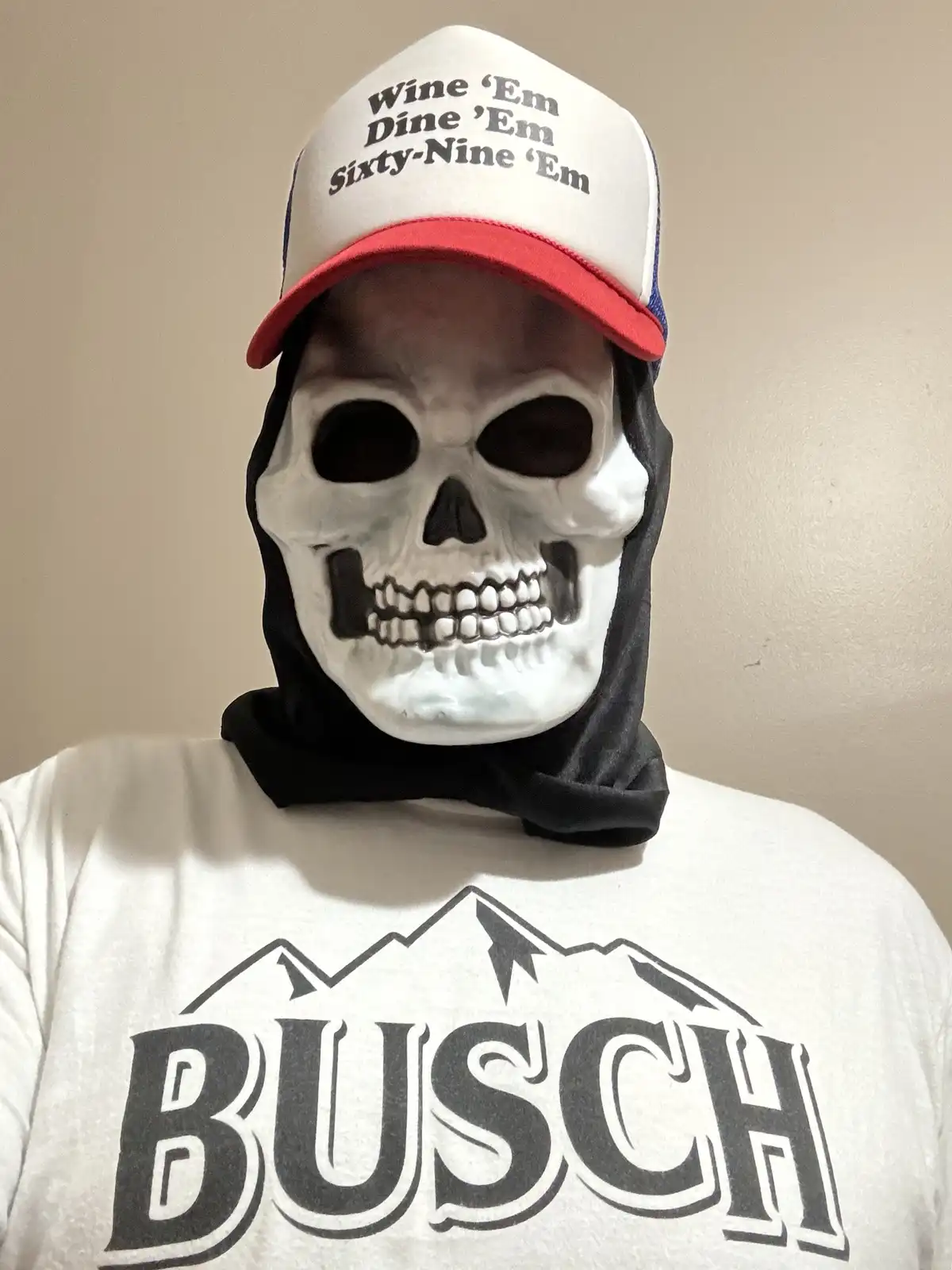 A person wearing a white skull mask over their face, a black neck gaiter, and a white t-shirt with a black 'BUSCH' logo and…