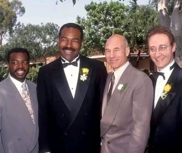 Four men in formal wear pose outdoors for a photo. From left to right: a Black man in a grey suit, a Black man in a black…