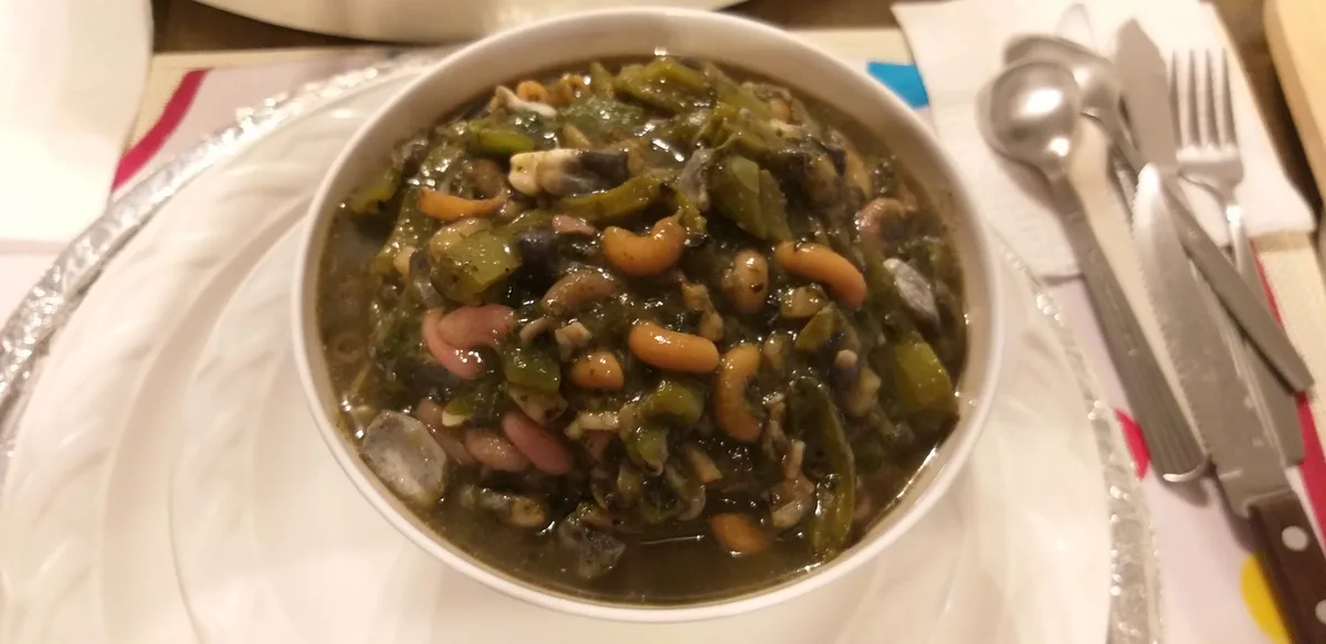 A close-up photo shows a white bowl filled with a dark green, thick stew containing various beans, chopped green peppers…