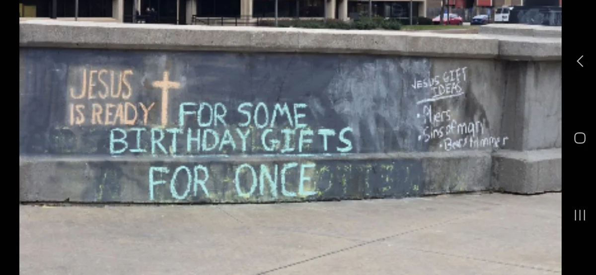 A concrete wall has chalk graffiti that reads 'JESUS IS READY FOR SOME BIRTHDAY GIFTS FOR ONCE' with a cross drawn next to…