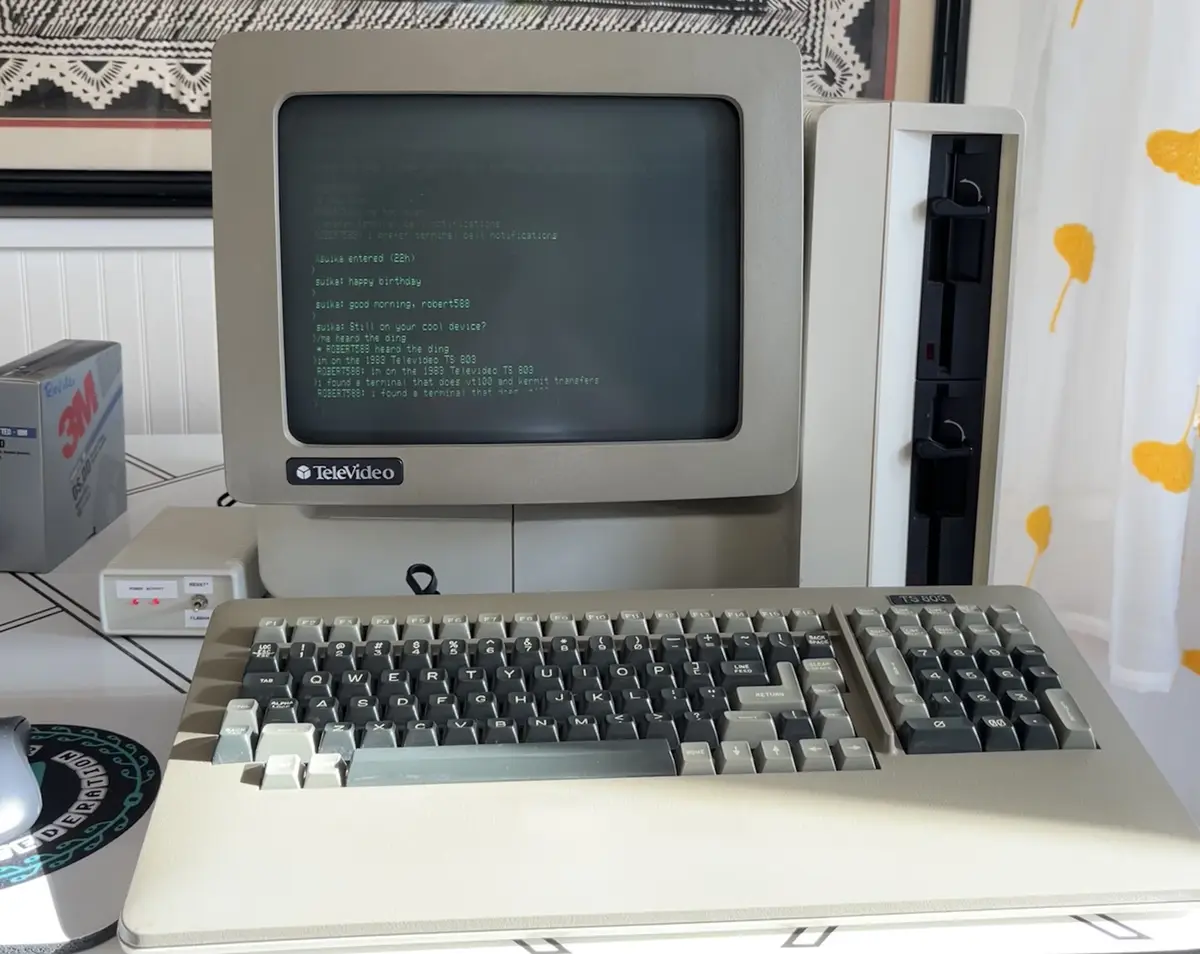 A vintage 1983 TeleVideo TS 803 terminal with a beige keyboard and monitor displays a green text chat log where user…